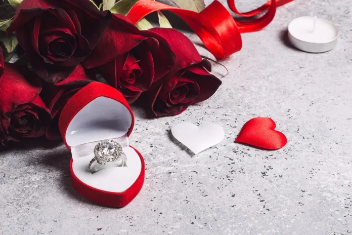 A diamond ring in a red heart-shaped box placed beside red roses and decorative heart shapes on a textured surface, styled as a romantic jewelry display.